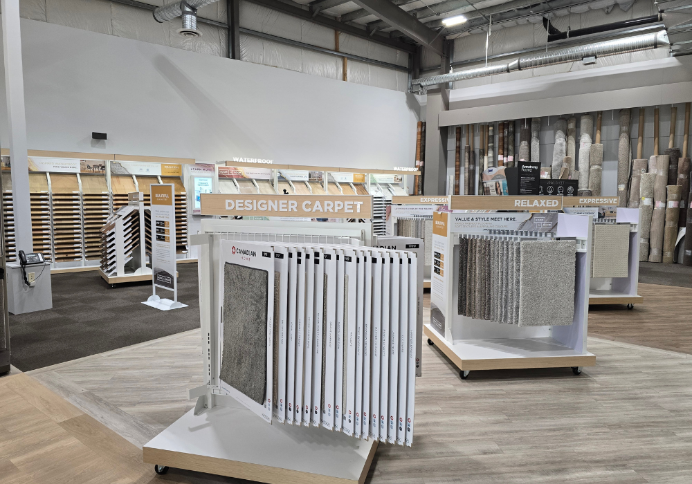 Kimball Carpet One showroom with wood flooring, a central display rack showing carpet samples, and surrounding shelves labeled “DESIGNER CARPET” and “RELAXED,” with rolls and swatches arranged along the walls.